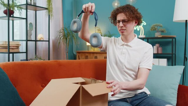 Unhappy young Caucasian man guy unpacking carton box, feeling dissatisfied with getting damaged wireless headphones order, writing negative feedback in mobile application, internet shopping concept.