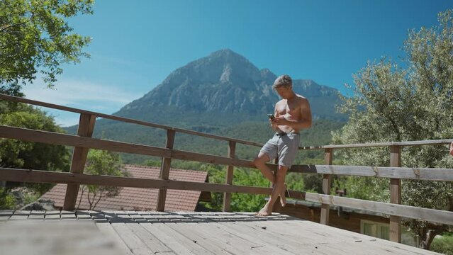 Adult shirtless man with fit athletic body enjoying warm sunny day on wooden veranda with majestic scenic mountains on background. smiling man using phone outdoors for taking photo and video.