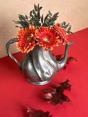 Vintage Metal Teapot with Vibrant Orange Gerbera Flowers and Autumn Leaves on Red Background.