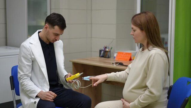 A male doctor in a white medical coat attaches a pulse oximeter sensor to the finger of a pregnant woman who is sitting opposite him on a chair in the emergency room. The doctor measures the patient's