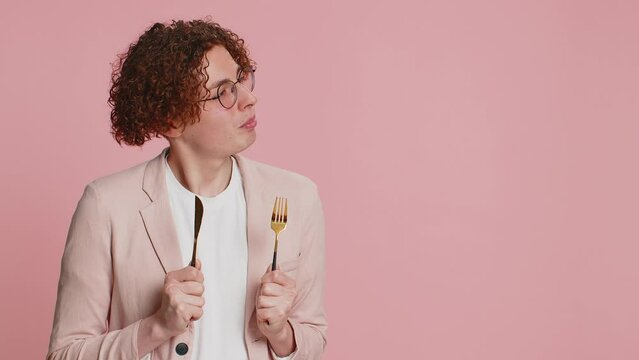 Ready to eat. Hungry Caucasian man with curly hair waiting for serving dinner dishes with with restlessness holding cutlery fork knife, will appreciate delicious restaurant meal on pink background