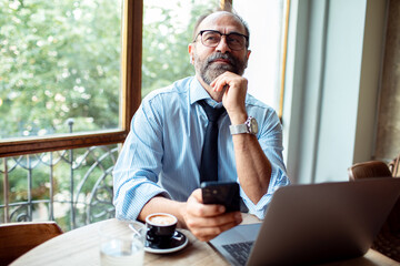 Thoughtful businessman holding phone and looking up in cafe