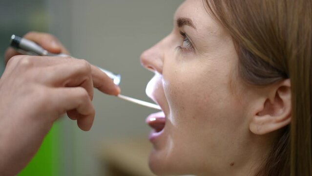 Close-up. A doctor asks a female patient to open mouth for a visual examination of the mucous membrane of the throat. The doctor uses a wooden spatula, to press the back of the tongue and a medical