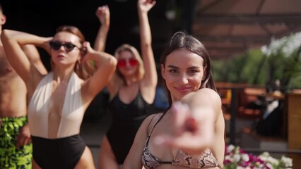Women and men posing, dancing and flirting on camera at summer pool party in colourful bikinis and sunglasses. Friends enjoying vacation at beach club resort with pool and sun loungers.