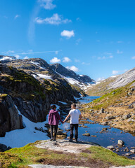 Naklejka premium A couple overlooking a serene mountain valley in Norway. The pristine blue sky and the lush green slopes, dotted with snow, create a breathtaking summer landscape. Kjeragbolten Norway