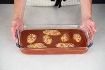 Female chef in an apron preparing meat or cutlets patties with rice, baking them in a dish with tomato sauce. Home cooking, traditional recipes, and culinary preparation.
