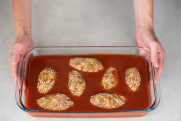 Female chef in an apron preparing meat or cutlets patties with rice, baking them in a dish with tomato sauce. Home cooking, traditional recipes, and culinary preparation.