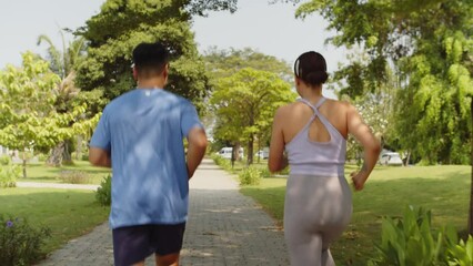Tracking shot of sporty couple in activewear running in park in summer morning, back view