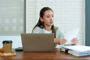 Beautiful young Asian female barista sitting at table in coffee shop Stressed about checking the store's accounts with laptop computer Drink coffee in afternoon to keep yourself from feeling sleepy.