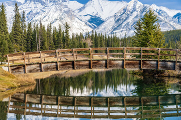 Wooden arch footbridge in Cascade Ponds park in autumn sunny day, snow-covered Mount Astley reflection on the water surface. Banff National Park, Canadian Rockies, Alberta, Canada.