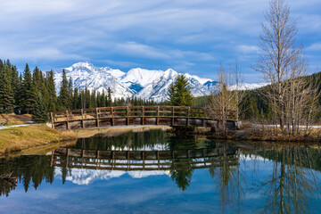 Fototapeta premium Wooden arch footbridge in Cascade Ponds park in autumn sunny day, snow-covered Mount Astley reflection on the water surface. Banff National Park, Canadian Rockies, Alberta, Canada.