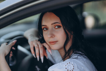 woman with black hair sits in the driver’s seat of a grey sedan car, facing the camera. The photo is a portrait, capturing her serene expression and the interior details of the vehicle