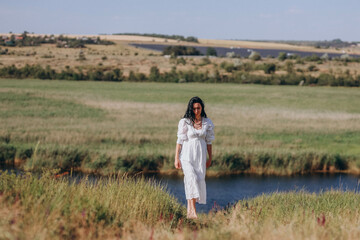 lady with black hair in a white long dress stands gracefully in a serene Ukrainian field by a calm river, embodying the natural beauty and tranquility of the countryside
