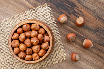 Overhead view of a bowl full of hazelnut kernels on a wooden surface. A piece of burlap and a few shelled hazelnuts lying around