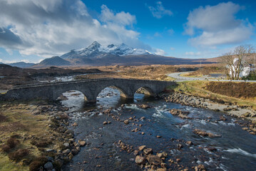 Old vintage brick bridge crossing river in Sligachan, Isle of Skye, Scotland with scottish landscape, vegetation, hills and mountains with fresh, blue water and sunny sky