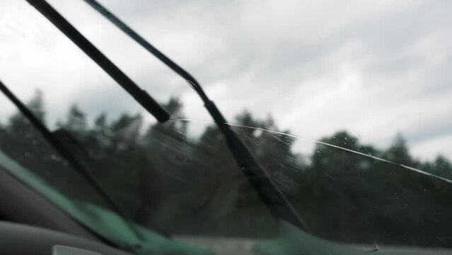 Close-up POV from inside a car onto raindrops on the windshield. Traveling on a wet road on a rainy day. First person view car passenger. Wipers wipe windshield from raindrops