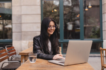 Student woman wear leather jacket, eyeglasses using laptop at cafe. Smiling young beautiful girl...