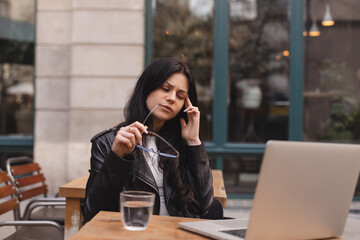 Tired student woman with laptop in outdoor cafe, woman touching her head with her glasses off, city street background. Headache, migraine or tired.