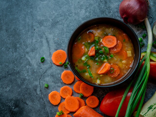 La imagen muestra un tazón de sopa con trozos visibles de verduras, como zanahorias y posiblemente algunas hojas verdes, sobre una superficie texturizada. Alrededor del tazón hay ingredientes.