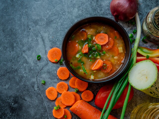 La imagen muestra un tazón de sopa con trozos visibles de verduras, como zanahorias y posiblemente algunas hojas verdes, sobre una superficie texturizada. Alrededor del tazón hay ingredientes.