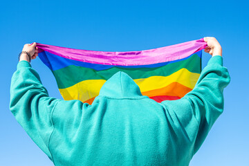 back view of a hooded person holding a flag with rainbow colors in support of the LGBTI community.