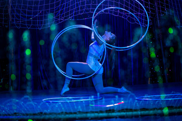 Young slim attractive circus gymnast woman performing live show with hula hoops on red arena with blue backlight, audience is excited