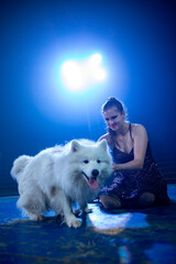 Young brunette caucasian woman artist on a blue circus arena after the show playing with big white dog Samoyed