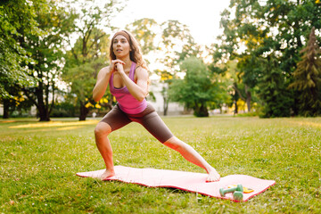 Young woman in sportswear stretching body on fitness mat at public park, doing yoga. Female taking...
