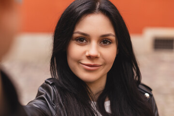 Close up portrait of young smiling woman tourist posing, walking in old town. Happy young model makes photo holding camera in hand close-up. Outdoor selfie photo in fashion outfit while walking.