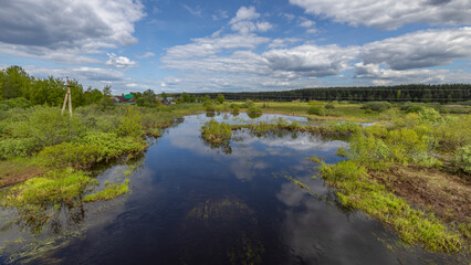 A wide shot of a winding river in a rural Russian countryside. The water is calm and reflects the blue sky and fluffy white clouds. Lush green vegetation lines the banks of the river