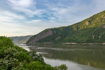  captures a serene river scene with a lush green forested cliff on the left. The calm water reflects the blue sky and scattered clouds, creating a tranquil and peaceful atmosphere.