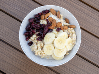 Top view of white bowl of oatmeal with sliced banana, almonds and dried cranberries on brown wooden table.