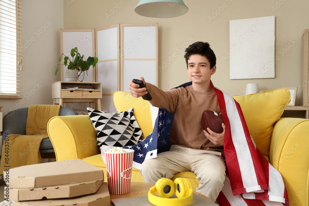 Wall mural Teenage boy with rugby ball and flag of USA watching football match at home