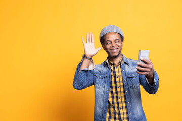 Carefree casual guy waves at webcam on videocall connection, talking to his family or friends in front of the camera. African american man in trendy outfit uses phone with online conference.