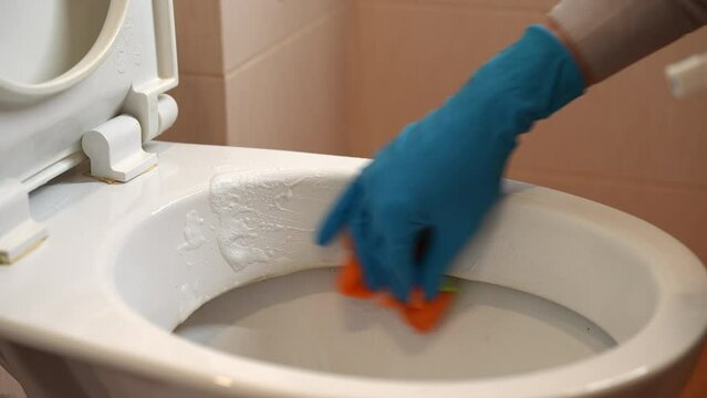 Close-up hands of unrecognizable househusband in gloves cleaning toilet bowl in bathroom. Housewife male doing cleaning and washing toilet bowl with sponge. Concept of domestic work, housekeeping.