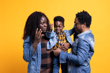 African american family of three smiling against yellow background, posing with affection and gratitude. Cheerful young parents holding their cute infant son, enjoying time together.