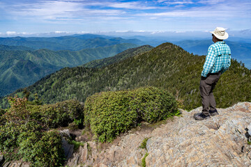 Naklejka premium A mature Caucasian man, a hiker, stands on a high rock looking at a scene of wooded mountain tops and ridges against a cloudy sky, Mt Le Conte, Great Smoky Mountain National Park, Tennessee