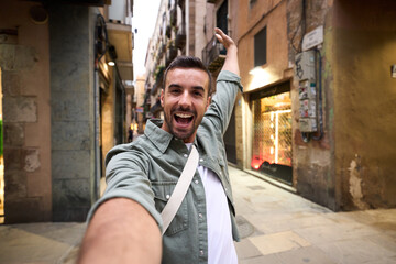 Young excited Caucasian man taking selfie on Paris touristic city street in background. Millennial male tourist takes happy picture of himself raising hand with joy and surprised gesture outdoors