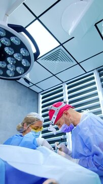 Two medics stand bent over the patient under surgery. Female scrub nurse assists the doctor performing operation on eye for hypothyroidism. Vertical video.