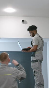 Two multi ethnic installers in uniform sets up security cameras in office corridor using laptop. Upset African American worker looks at camera. Concept of surveillance systems and privacy. Quick zoom