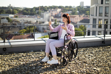 Young woman in wheelchair on rooftop taking a selfie with smartphone. She is smiling and enjoying the cityscape around her. The scene portrays independence, accessibility, and modern urban living.