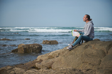 Man sitting on a rock by the ocean holding a map, exploring the coastline