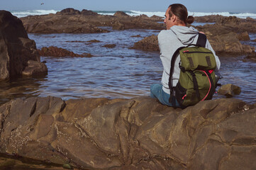 Fototapeta premium A man wearing a gray hoodie and green backpack sits on a rocky shore, looking out at the tranquil sea. The scene evokes a sense of solitude, reflection, and connection with nature.