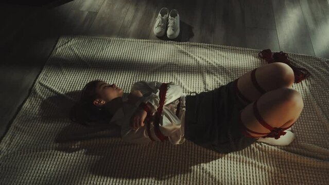 Woman lying on rug practicing art of Shibari with red ropes. Theme of restraint and vulnerability. White sneakers placed aside. Concept of artistic expression and tradition