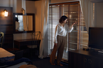 Worried businesswoman looking through blinds while standing in hotel room