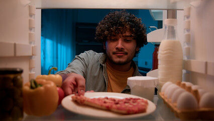 View inside from refrigerator Indian sly man with curly hair open fridge at night evening in kitchen hungry guy male looking around take out slice of pizza overeating at nighttime unhealthy nutrition