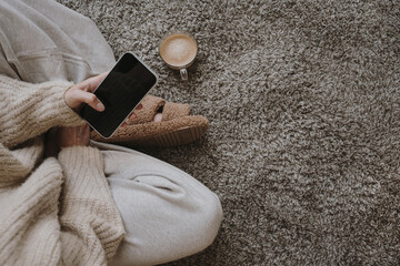 Woman sitting on fluffy carpet with cup of coffee and using mobile phone. Cozy morning routine...
