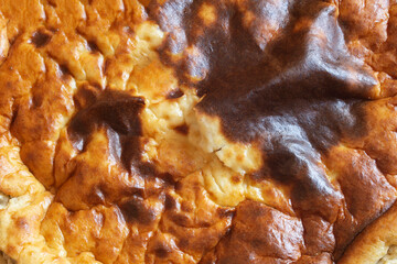 Close-up of a baked cheesecake with a golden-brown crust that has some darker brown spots with an uneven texture with cracks and crevices, suggesting it is crispy on the outside. Abstract background.