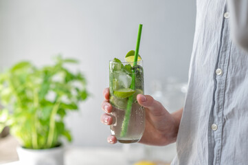 Person hand holding glass with mojito cocktail drink with ice, mint, lime and green drinking straw on neutral gray wall background, summer refreshing beverage, soft natural sunlight, lifestyle