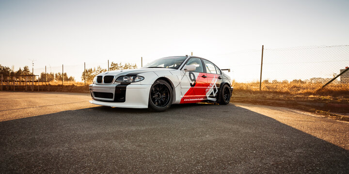 white-red-black BMW E46 racecar near the fence and dry grass. Three quarter front view of widebody sedan at sunset. Kyiv region, Ukraine - March 16 2015.